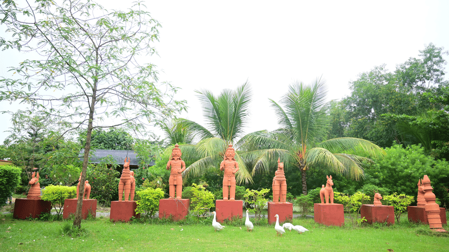 An outdoor area, featuring a green lawn and several terracotta statues or planters shaped like people and animals, set against a backdrop of trees at Pipul Odi Art Museum Resort.