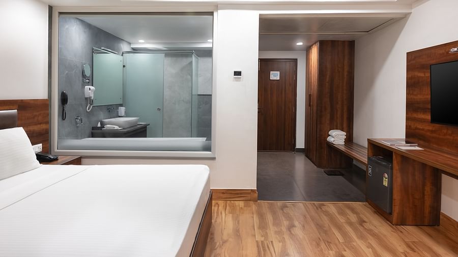 A modern hotel room with a large bed, wooden flooring, a TV, and a glass-walled bathroom in view.- Clarks Inn Express, Near Jammu Bari Brahmana Railway Station