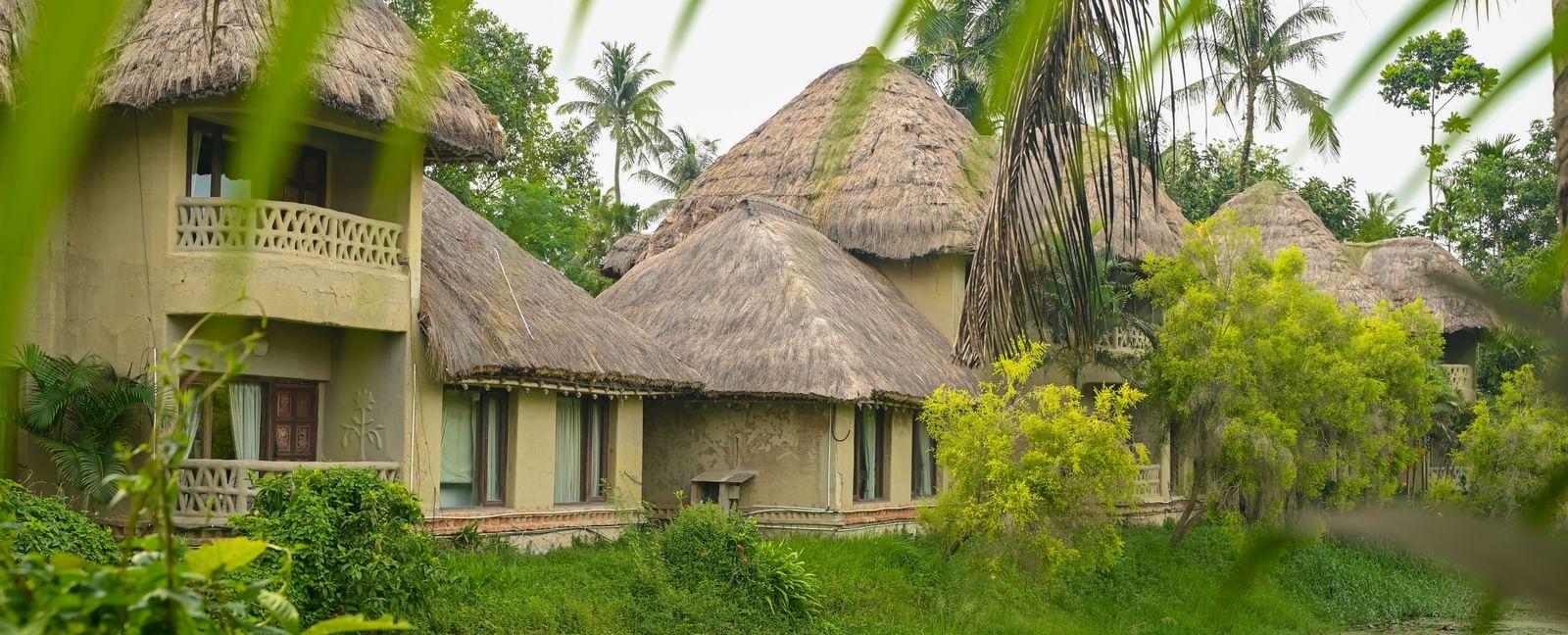 Thatched-roof Earth Villas at Vedic Village Spa Resort overlooking a water body, with lush green vegetation lining the perimeter of the natural surroundings.