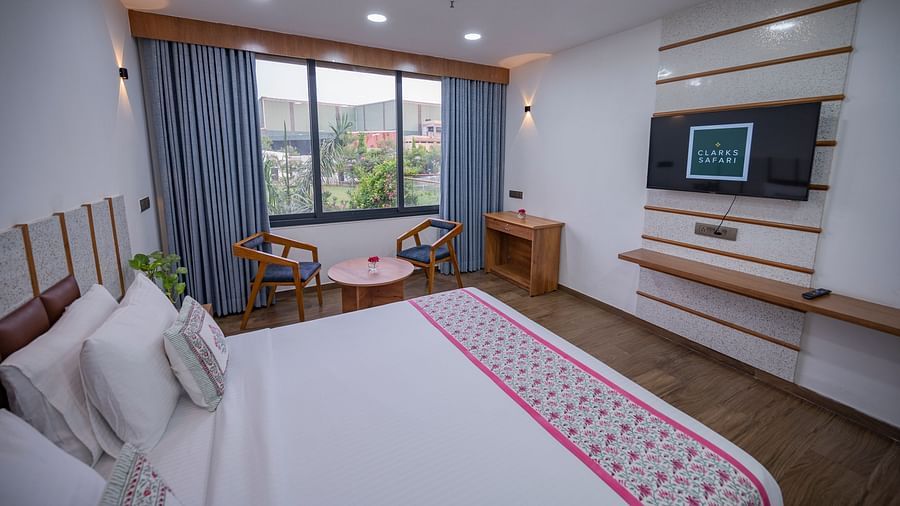 The view of a double bedroom at Clarks Safari Kheda with wooden flooring, large window with curtains, tv, two chairs, round table, desk, remote, flower vase on top of the table and neatly arranged pillows.
