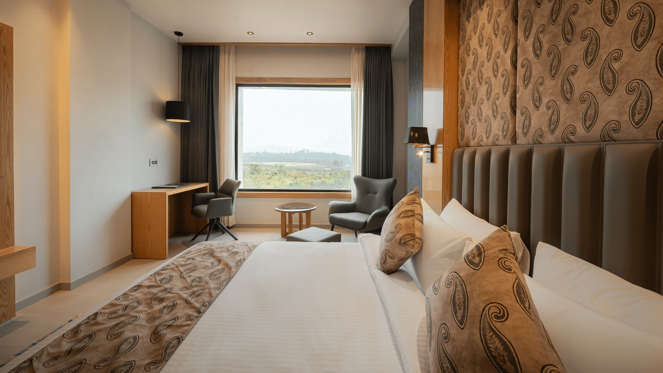 A bright guest room with a king-sized bed, a patterned headboard, and a large window overlooking the hills at Ananta Express, Katra.