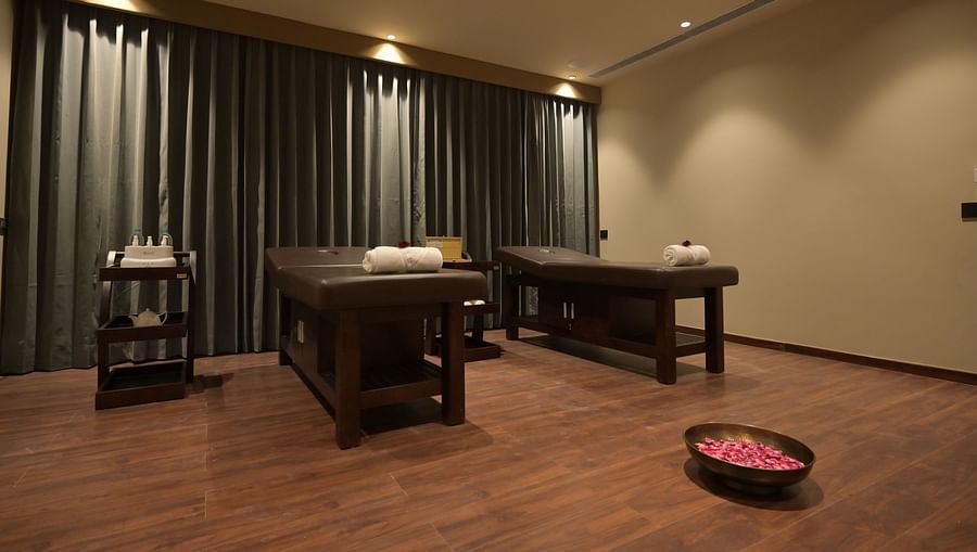 A softly lit spa treatment room features two massage tables with rolled towels, dark curtains, and a bowl of rose petals on the floor. - Clarks Safari Haridwar