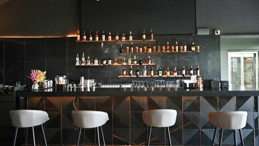 Modern bar at Hotel Daspalla, Hyderabad with black marble backdrop, premium spirit display on lit shelves, white bar stools, and curved wooden ceiling beams with warm lighting.