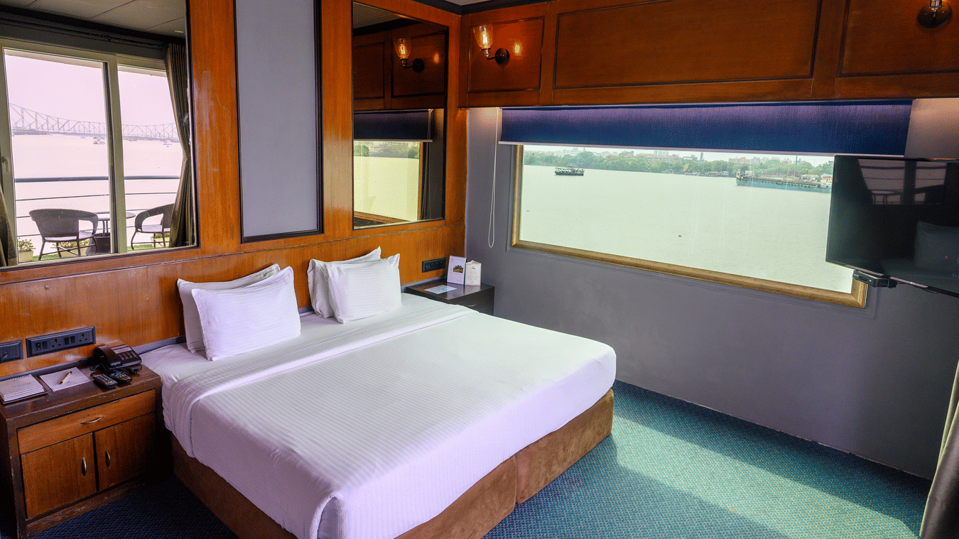 A hotel bedroom with a large bed and a wide window offering a view of the river. Wooden paneling and a TV are visible.