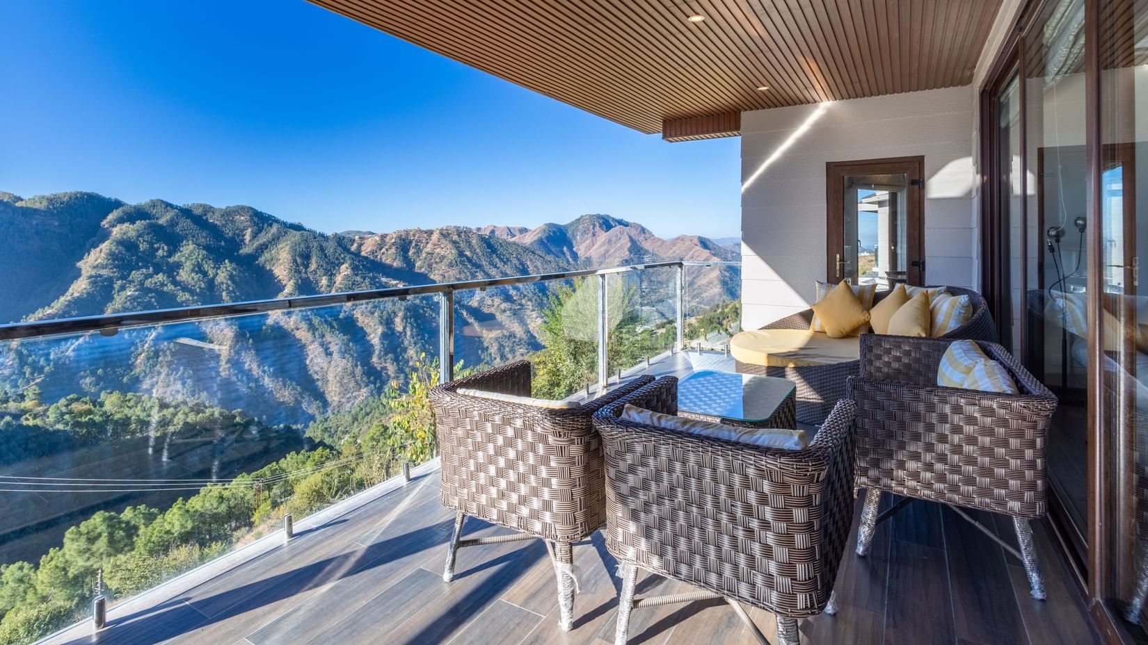Balcony of The Manor Luxury Apartments, Shimla, featuring wicker chairs and a table overlooking a stunning mountain valley and blue sky views.