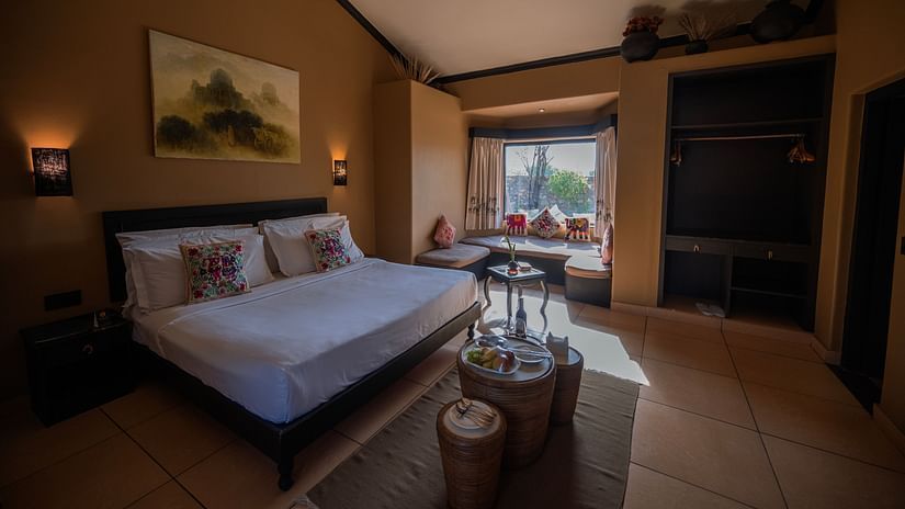 Interior of a hotel room showcasing a double bed, brown walls, a painting, and a window seat area with pillows and a table - Clarks Safari, Pushkar