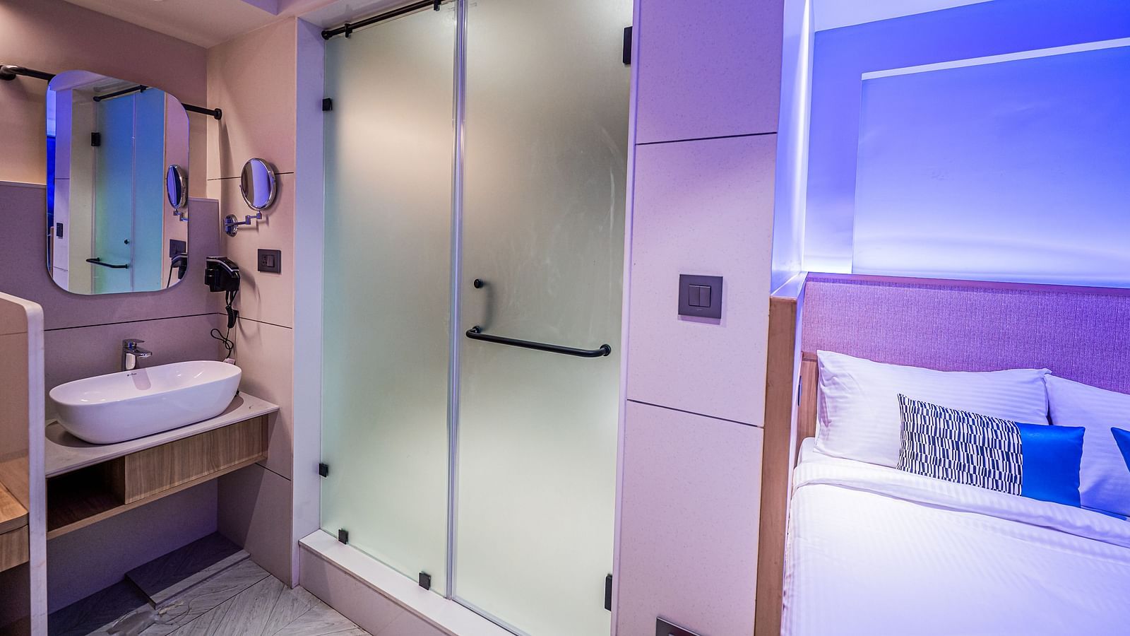 A washbasin below a wall-mounted mirror with a tinted door beside it, and a bed placed next to it at Indie Stays Mumbai International Airport.