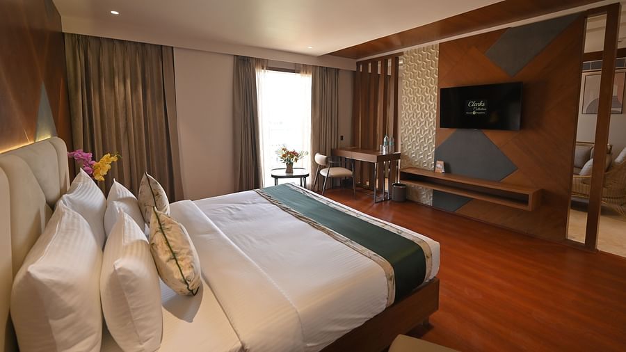 Side view of the bedroom featuring a wall-mounted television and a window with partially drawn curtains at Clarks Collection Grand Sapphire, Ajmer.