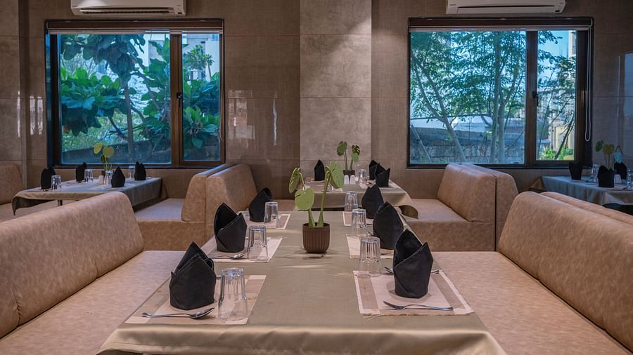 Dining table set with black tissue,spoons, flower vase beside large windows, ac and cushioned seating at Clarks Collection Somnath.