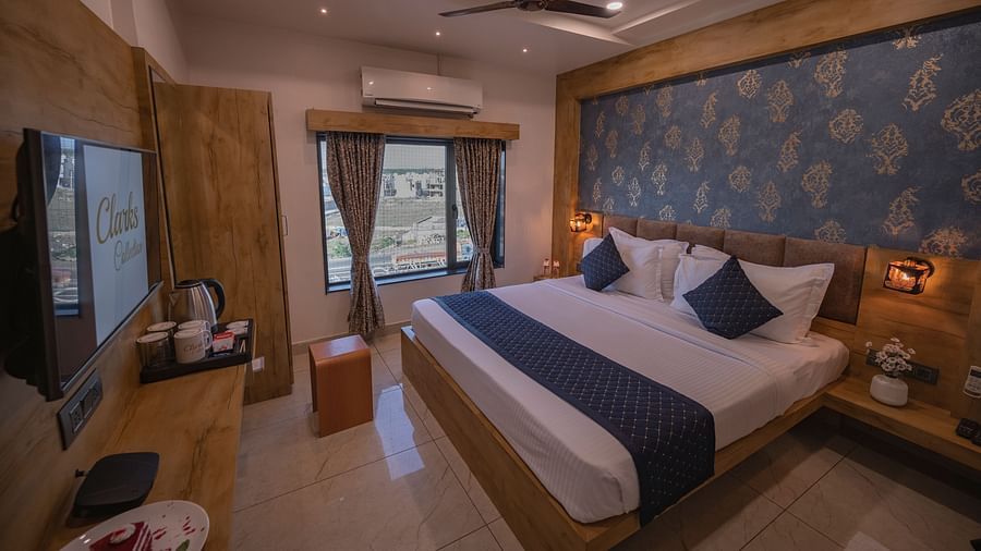 A bedroom with wooden interiors, double bed, ac, water bottles, bedside table with flower vase on the top, tv, and twin lamps at Clarks Collection, Somnath.