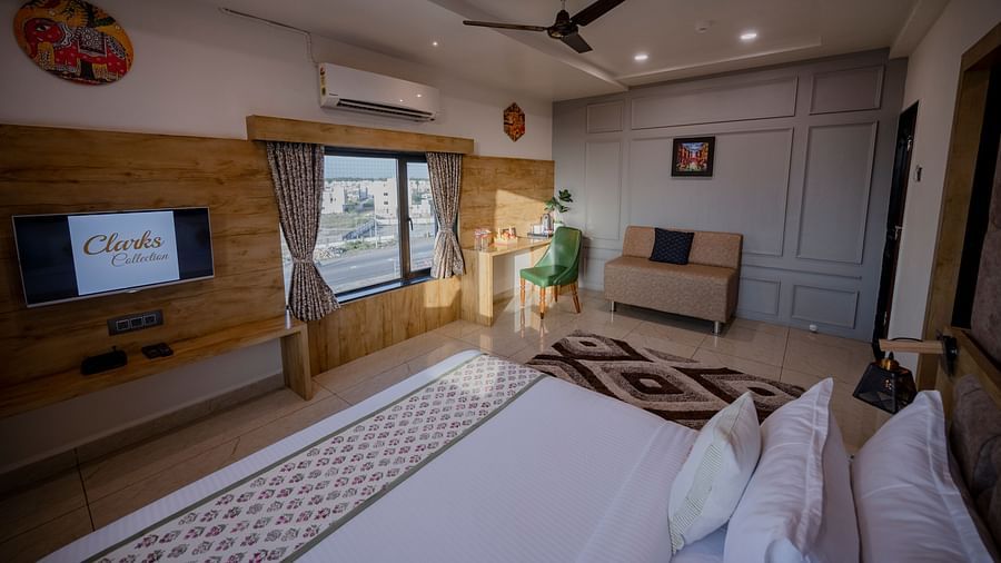 Side view of a Clarks Collection Somnath bedroom with tv, bed, fan, rug, wooden decor, and natural light from windows.