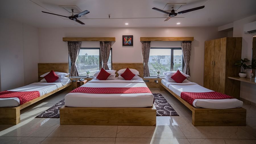 Three-bed setup at Clarks Collection Somnath with red accents, windows with curtains, ceiling fans, and a wall-mounted photo frame.