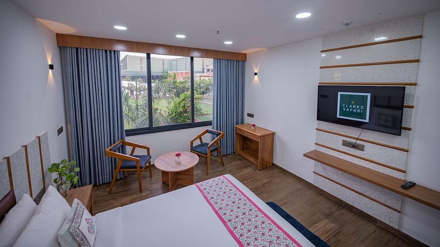 The view of a double bedroom at Clarks Safari Kheda with wooden flooring, large window with curtains, tv, two chairs, round table, desk, remote, flower vase on top of the table and neatly arranged pillows.