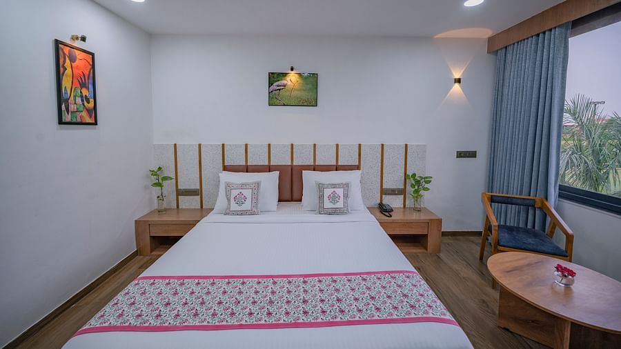 A bedroom at Clarks Safari Kheda with double bed, artwork on walls, curtains open to outside view, bedside table with plant on top, round table with flower vase on top, and wooden side furniture.