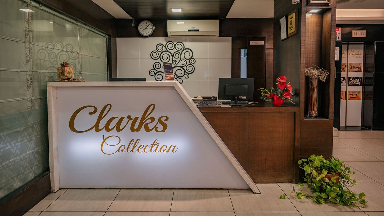 Reception area at Clarks Collection, Vadodara, featuring a white front desk with signage of Clarks Collection, a wall clock, desktop monitors, and indoor decorative plants.