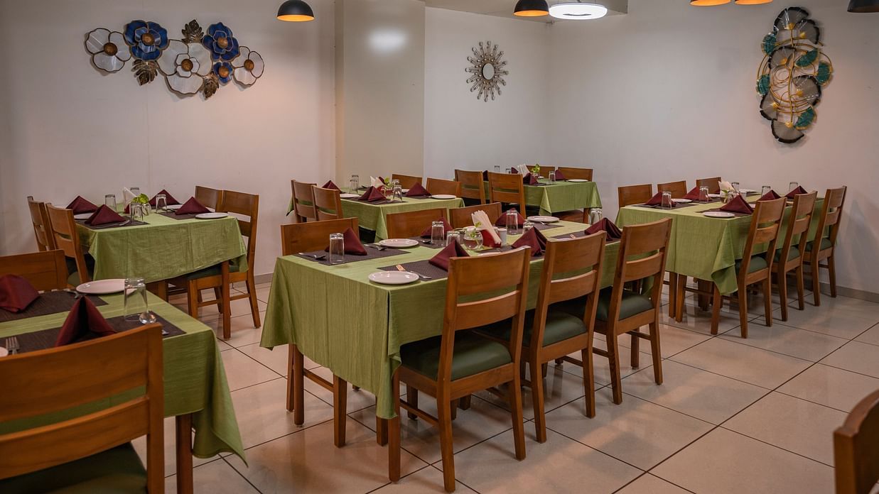 Clarks Safari Kheda's dining area with multiple tables are set with neat coverings and chairs, lights, neatly arranged tissues, plates, spoons, a small money plant on the centre and decorative elements on the walls.