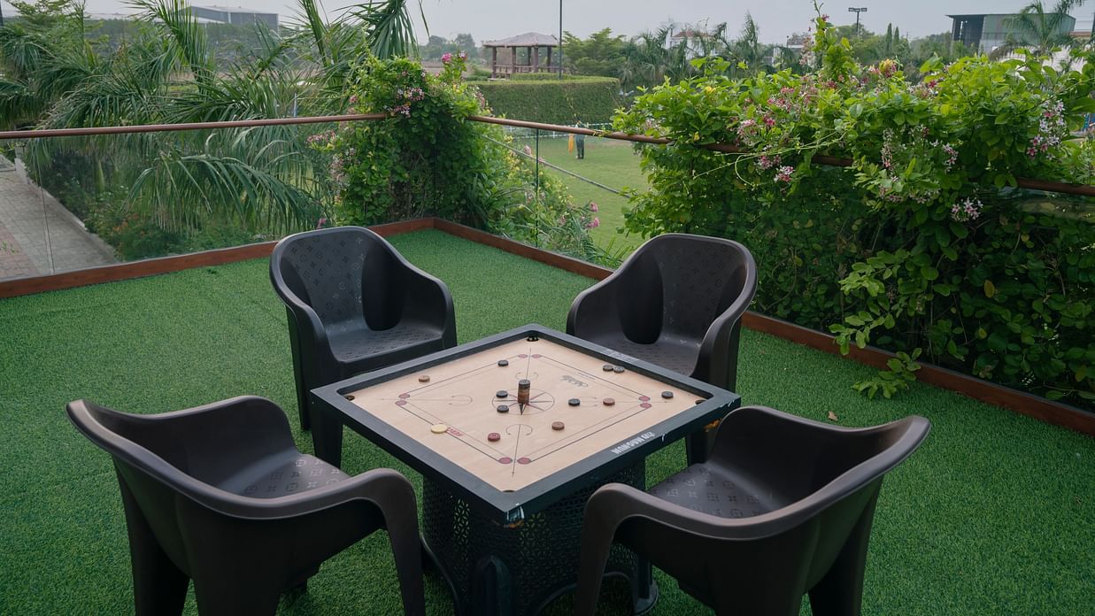 An outdoor seating area at Clarks Safari Kheda with a table, four chairs, and a board game set up, on a low,  surrounded by green surface and trees.