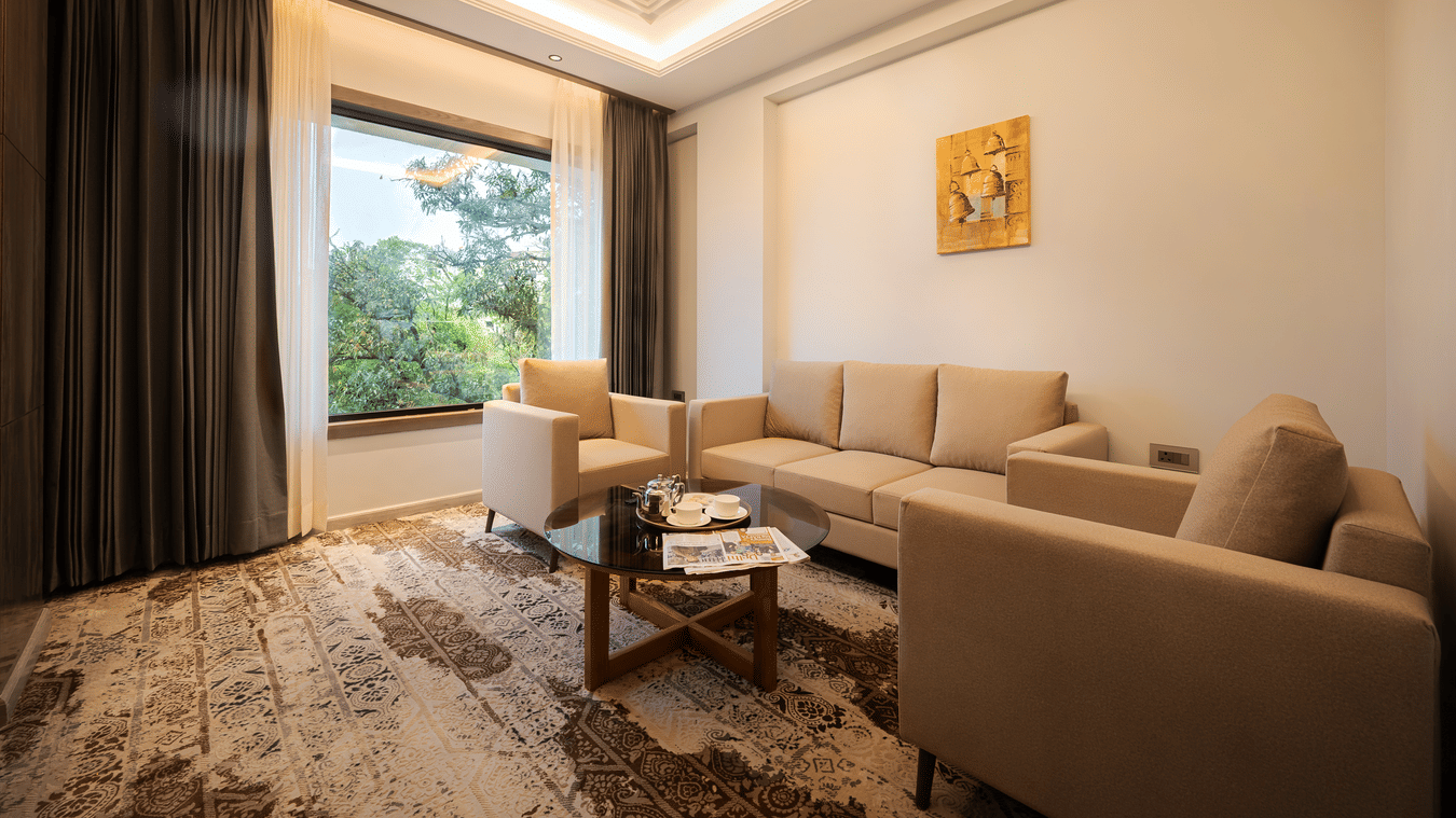 A bright suite living area with a large beige sofa, a coffee table, and a window with grey curtains at Ananta Express, Katra.