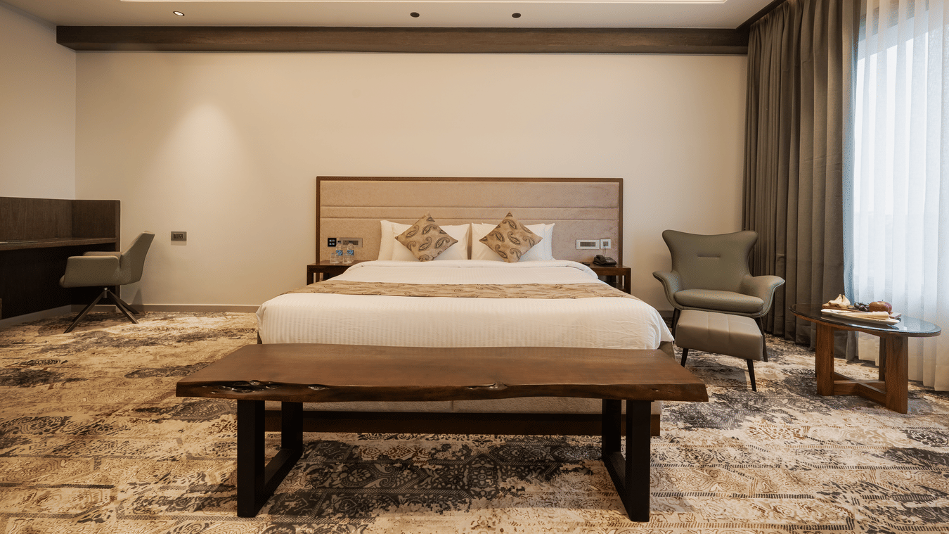 A guest room featuring a king-sized bed, a wooden bench, and a work desk area with an armchair at Ananta Express, Katra.