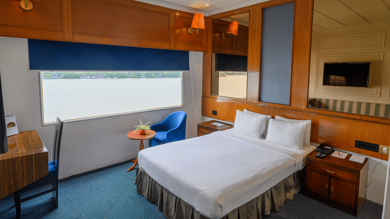 Polo Floatel Kolkata bedroom with a large bed, blue chair, and desk, offering a view of the water from the window.