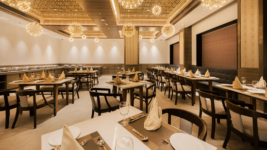 A spacious restaurant dining hall featuring wooden tables, black chairs, and modern gold-tone chandeliers at Ananta Express, Katra.