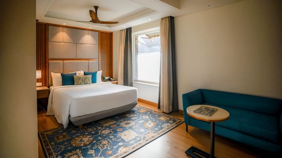 A brightly lit hotel room features a neatly made bed with white linens, a teal armchair, and a small round side table with a lamp. A window with sheer curtains allows natural light to filter in - Ananta Spa and Resort, Pushkar