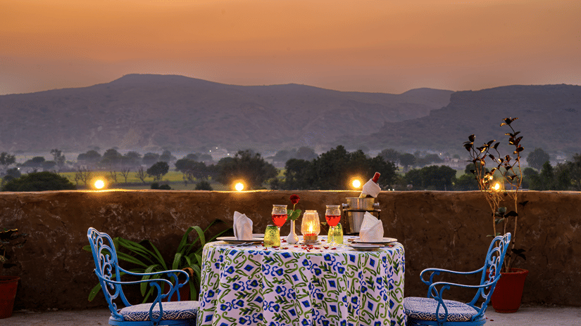 A blue patterned outdoor dining table set with drinks and napkins on an open-air terrace overlooking hills at sunset -  Hill Fort-Kesroli