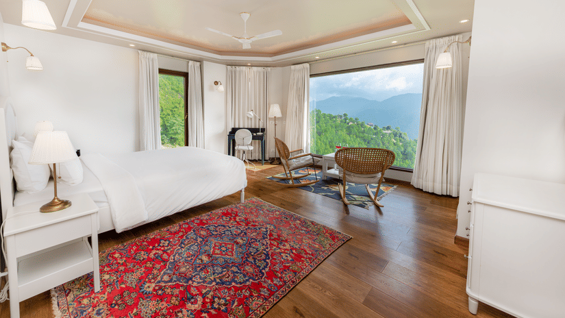 Bright bedroom at The Manor Luxury Apartments, Shimla, featuring a plushy bed, elegant furnishings, large picture windows, and panoramic views of the surrounding green hills.