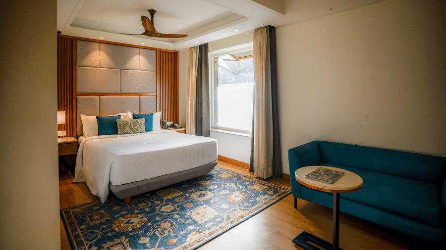 A brightly lit hotel room features a neatly made bed with white linens, a teal armchair, and a small round side table with a lamp. A window with sheer curtains allows natural light to filter in - Ananta Spa and Resort, Pushkar
