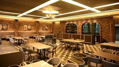 Dining area with multiple tables and chairs, patterned floor, and textured walls, lit by ceiling lights and a chandelier.