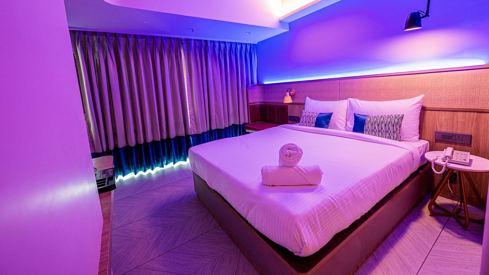 A spacious room at Indie Stays Mumbai International Airport features a large bed with neatly rolled towels, a window with closed curtains, and the room lit with cool lighting.