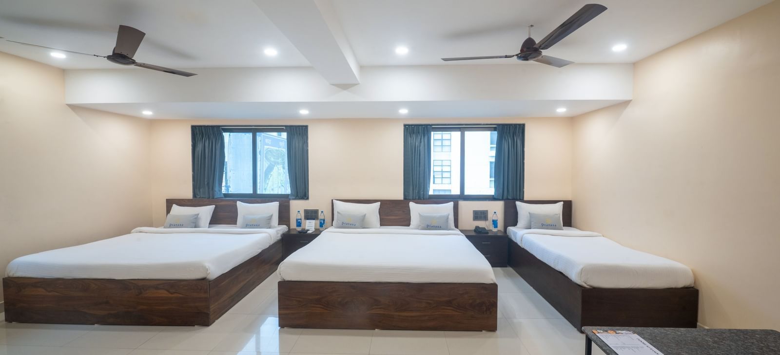 A large room at Pravasa Stay, Shirdi, contains three beds, two ceiling fans, and multiple windows.