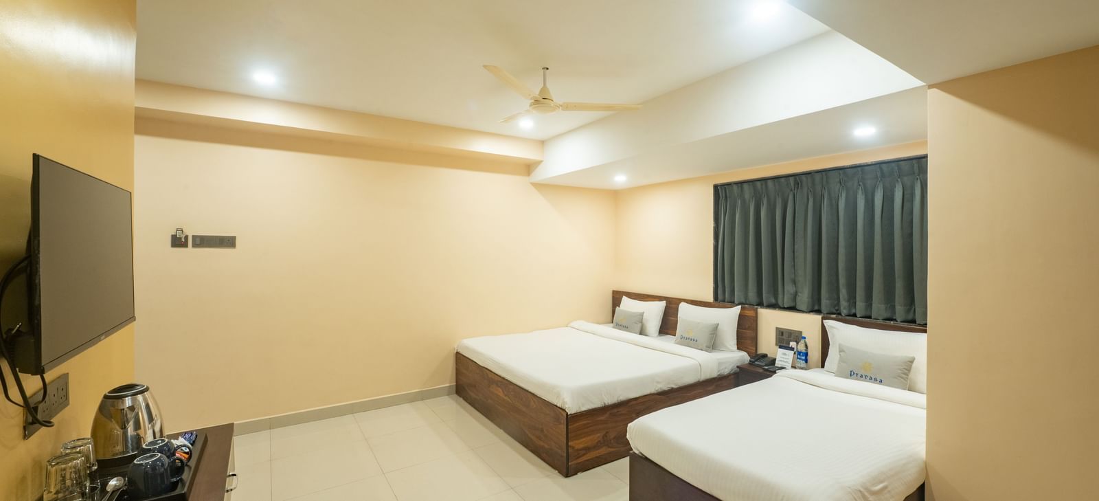 A room at Pravasa Stay, Shirdi, is equipped with two beds, a wall-mounted television, and dark curtains over a window.