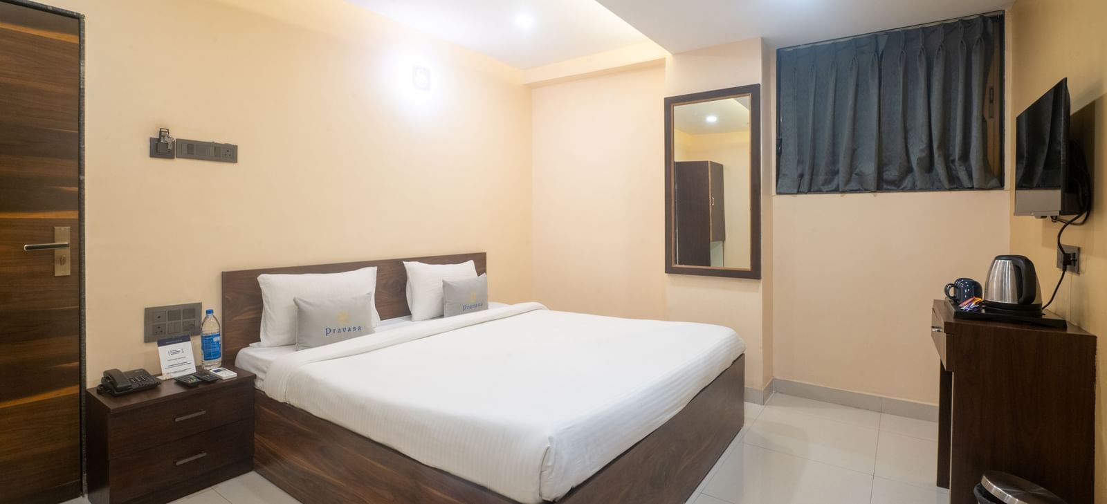 A room at Pravasa Stay, Shirdi, is furnished with a wooden bed, a bedside table, and a wall-mounted mirror.
