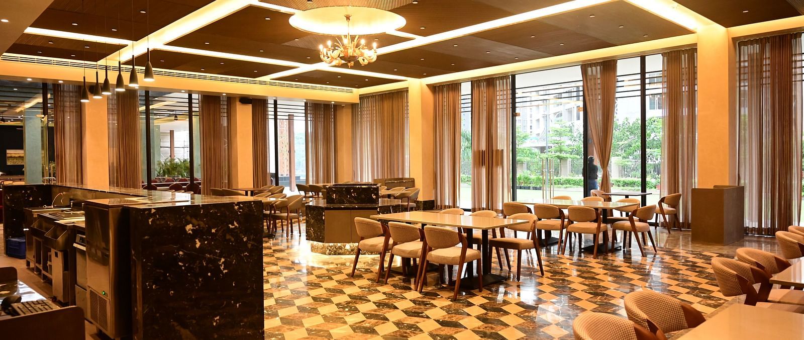 Dining area with tables, chairs, patterned floor, and large windows, lit by ceiling lights and a chandelier.