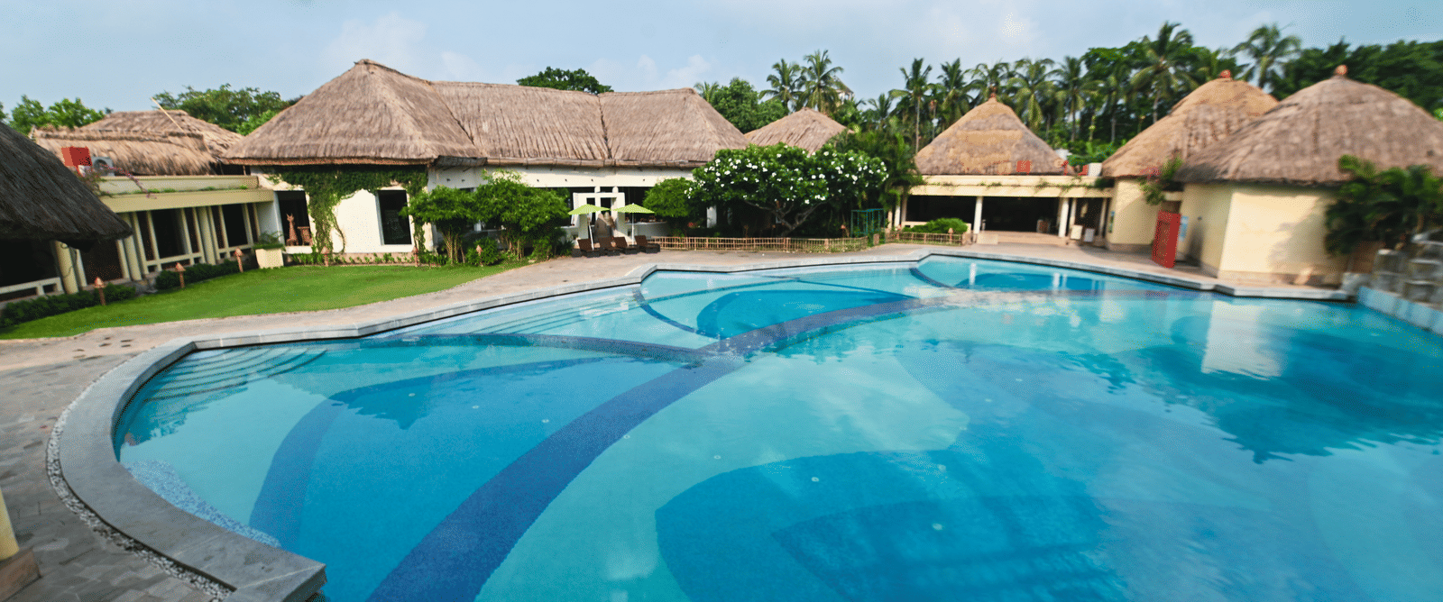 A daylight view of the common swimming pool at Vedic Village Spa Resort, featuring buildings with tatched roofs in the background.