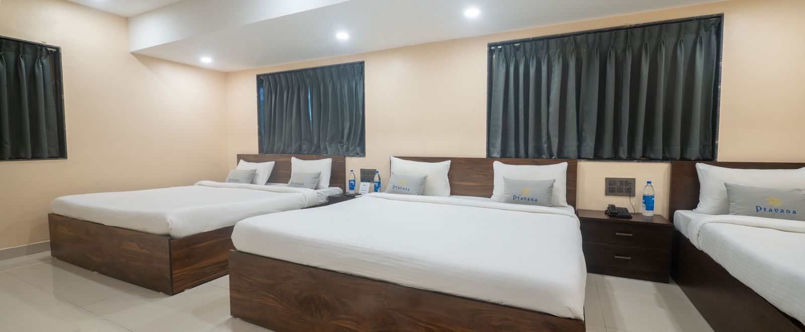 A large room at Pravasa Stay, Shirdi, contains three wooden platform beds with white linen and dark curtains.