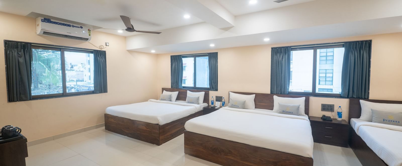 A large room at Pravasa Stay, Shirdi, features three beds, two windows, and two dark ceiling fans overhead.