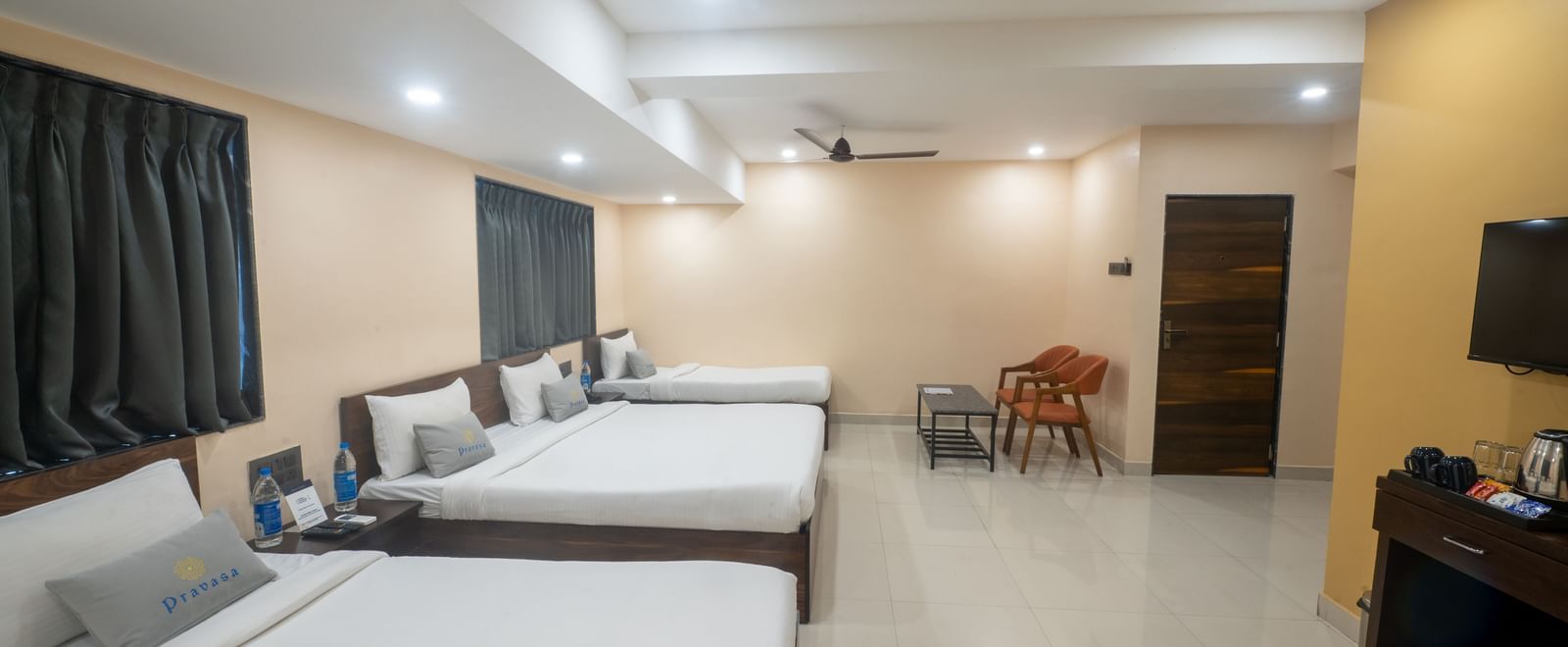 A large room at Pravasa Stay, Shirdi, features three beds, two ceiling fans, and a small sitting area.