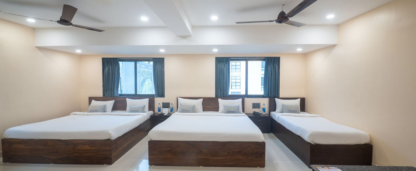 A large room at Pravasa Stay, Shirdi, contains three beds, two ceiling fans, and multiple windows.
