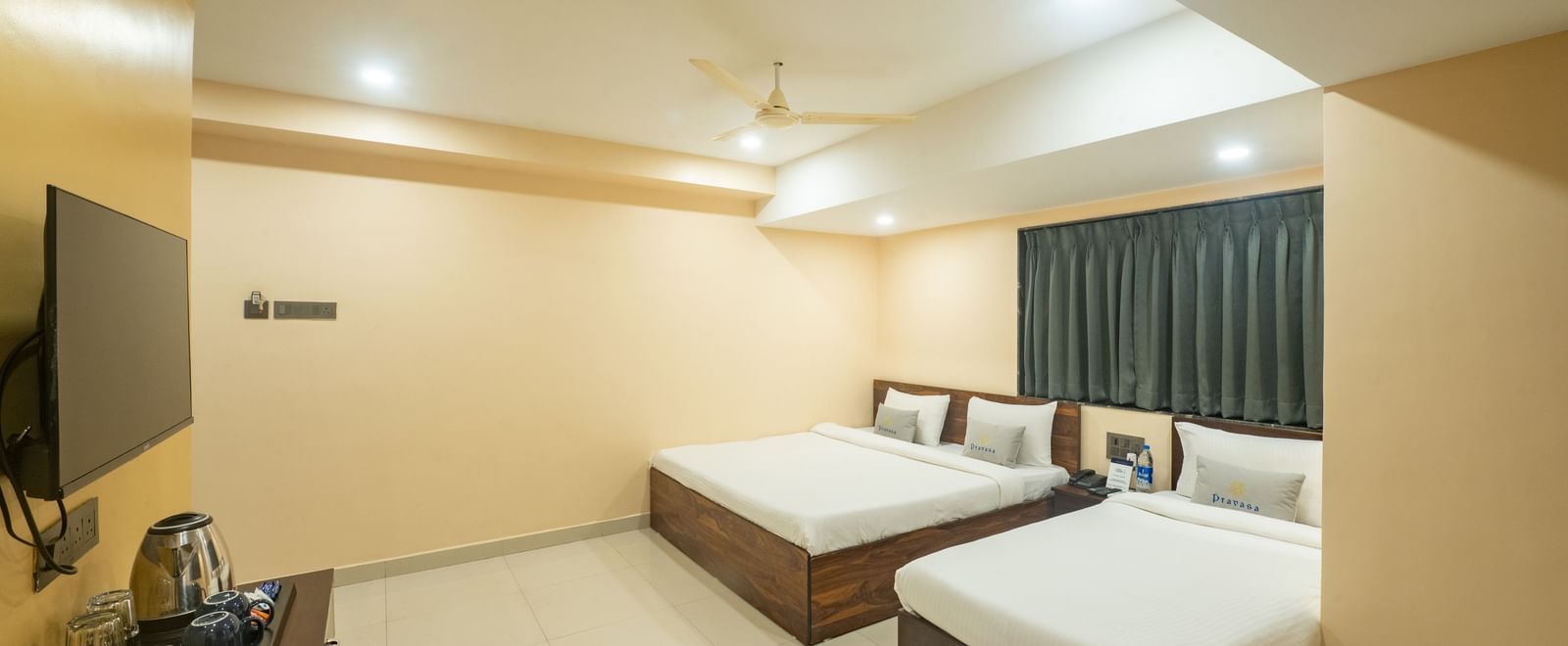 A room at Pravasa Stay, Shirdi, is equipped with two beds, a wall-mounted television, and dark curtains over a window.