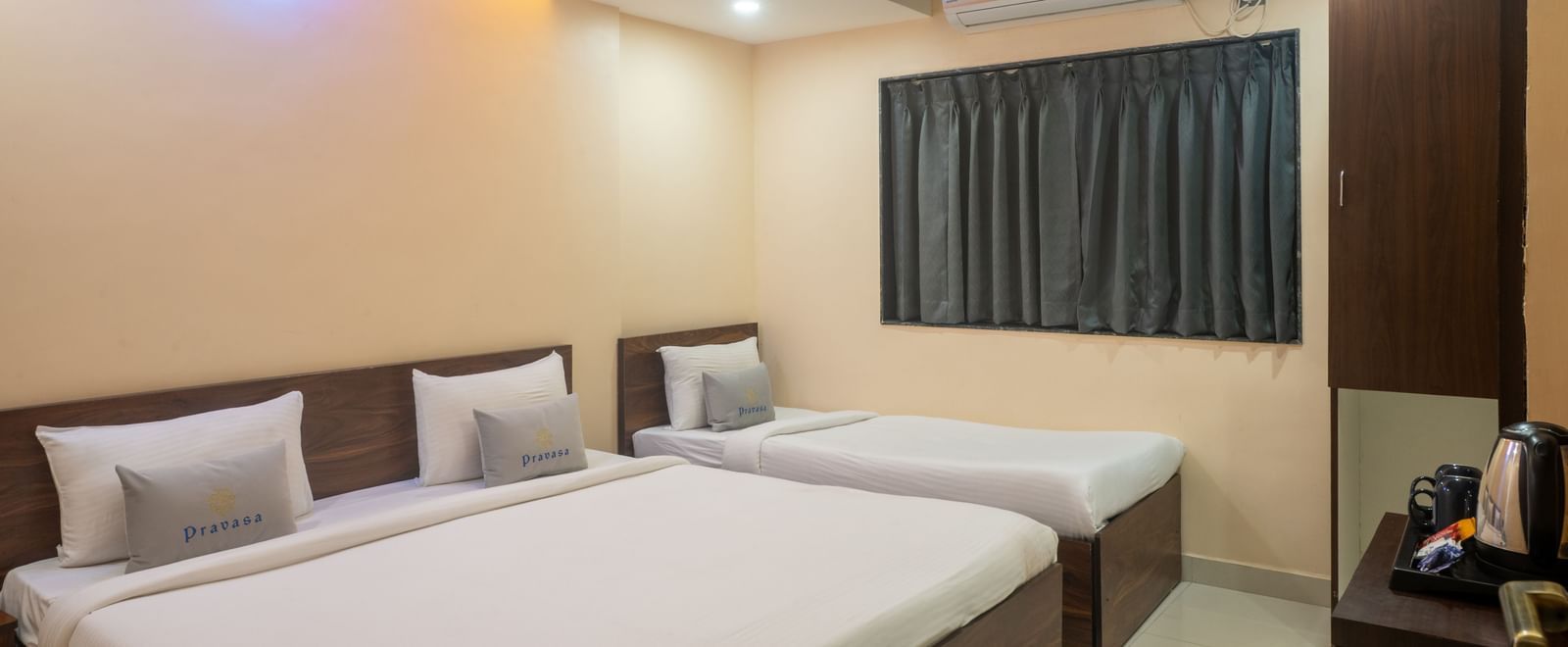 A room at Pravasa Stay, Shirdi, is equipped with one double bed and one single bed with a dark wardrobe nearby.