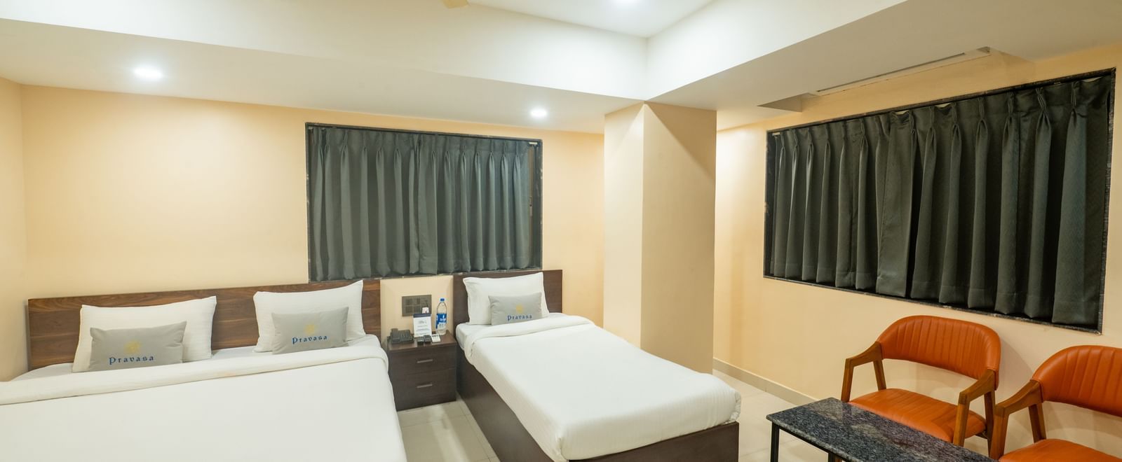 A room at Pravasa Stay, Shirdi, has two beds, two orange chairs, a small table, and a ceiling fan.