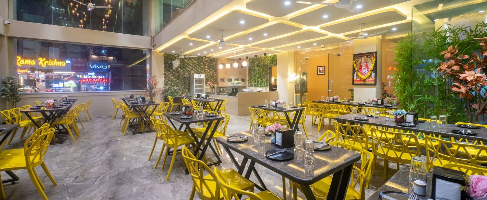 The dining space at Pravasa Stay, Shirdi, features bright yellow chairs and many ceiling lights.