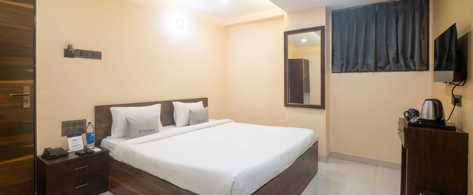 A room at Pravasa Stay, Shirdi, is furnished with a wooden bed, a bedside table, and a wall-mounted mirror.