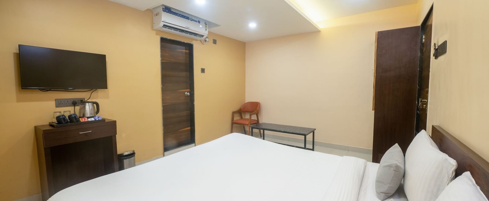 A room at Pravasa Stay, Shirdi, features a large bed with white linen, a desk, and a wall-mounted television.