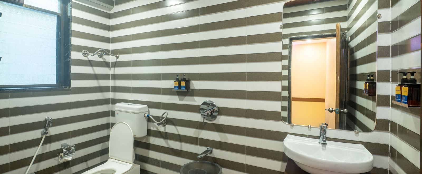 A bathroom at Pravasa Stay, Shirdi, features striped wall tiling, a modern toilet, and a washbasin with a mirror.