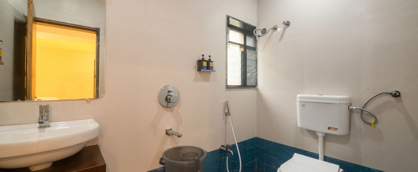 The bathroom at Pravasa Stay, Shirdi, features a washbasin on a wooden vanity, a toilet, and shower fixtures.
