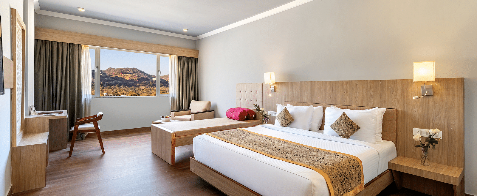 A bright hotel room with a neatly made bed, wooden furniture and a large window offering a view, with a minimalist design - Via Lakhela Resort & Spa, Kumbhalgarh