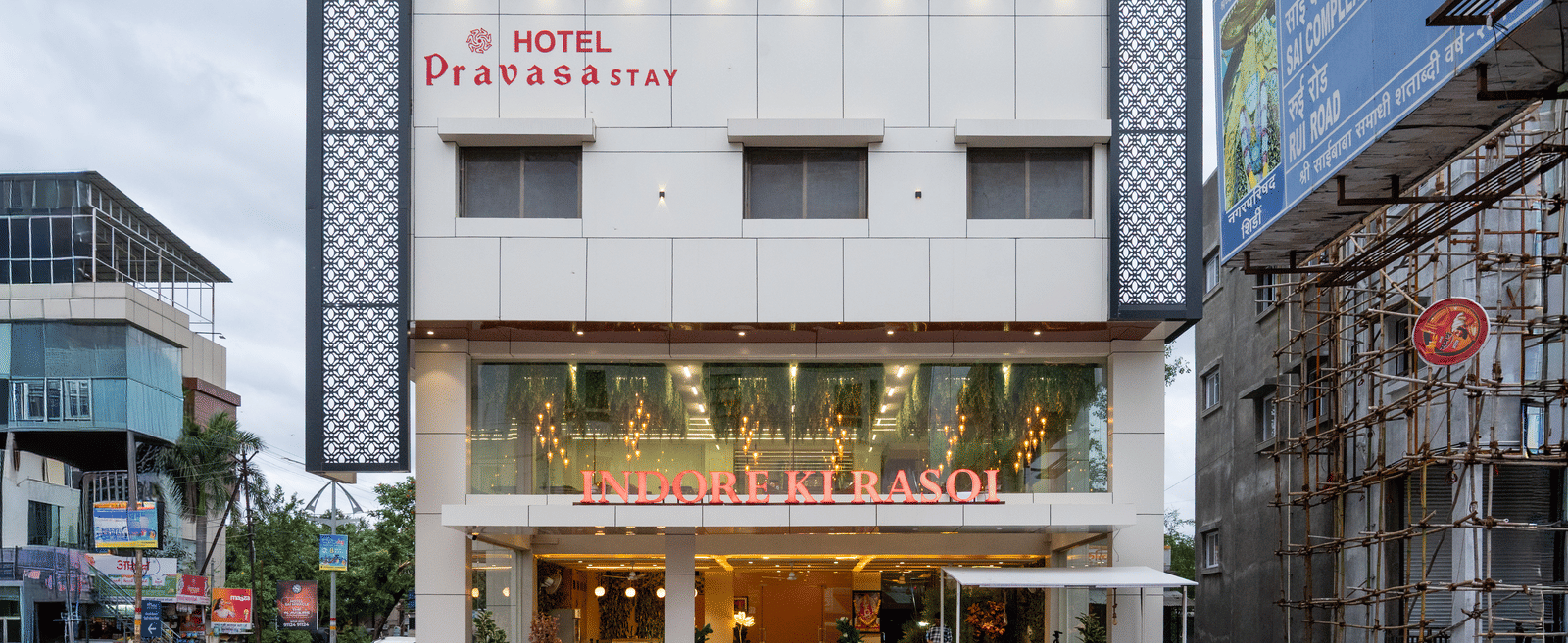 The main facade of Pravasa Stay, Shirdi, a white three-storey building, has a ground floor entrance and windows.